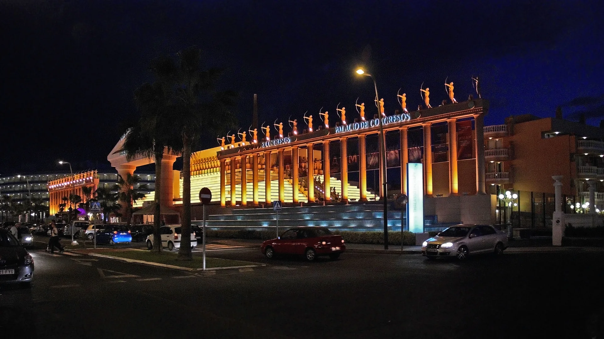Oświetlona kolumnada Palacio de Congresos w Playa de las Américas nocą, ze złotymi kolumnami, posągami łuczników wzdłuż dachu i głęboko błękitnym niebem nad nimi